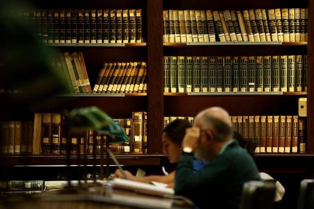 Una sala de lectura con personas leyendo sobre el escritorio y de fondo una estantería completa de libros.