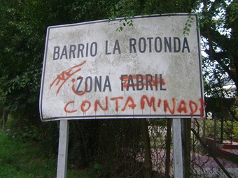 Cartel con el nombre del barrio y tachadura  donde dice zona urbana y agregado con  aerosol zona contaminada
