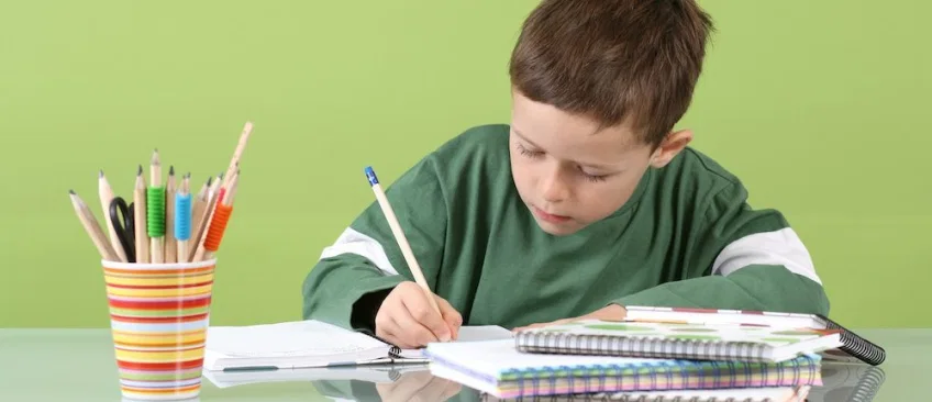 foto de niño de primaria estudiando en una mesa, con cuaderno y lápices. 