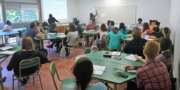 Fotografía del aula, lxs asistentes al seminarios distribuidxs en mesas redondas en grupos, una docente escribiendo en la pizarra y sobre pared lateral la presentación con diapositivas.